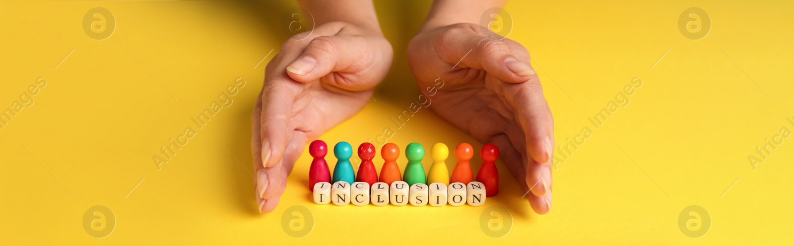 Image of Woman protecting colorful pawns and wooden cubes with word Inclusion on yellow background, closeup. Banner design