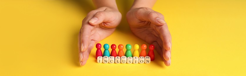 Woman protecting colorful pawns and wooden cubes with word Inclusion on yellow background, closeup. Banner design