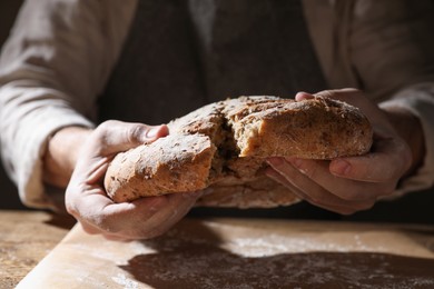 Photo of Man breaking loaf of fresh bread at wooden table, closeup