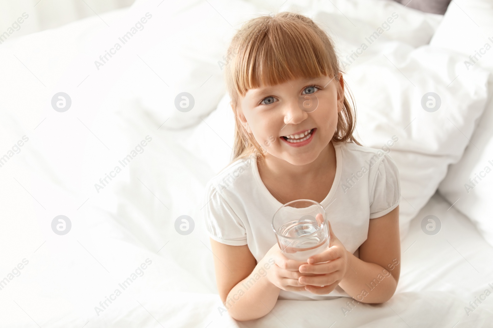 Photo of Cute little girl with glass of fresh water sitting in bed at home. Space for text