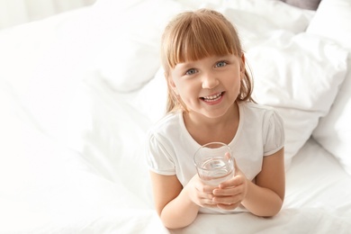 Photo of Cute little girl with glass of fresh water sitting in bed at home. Space for text