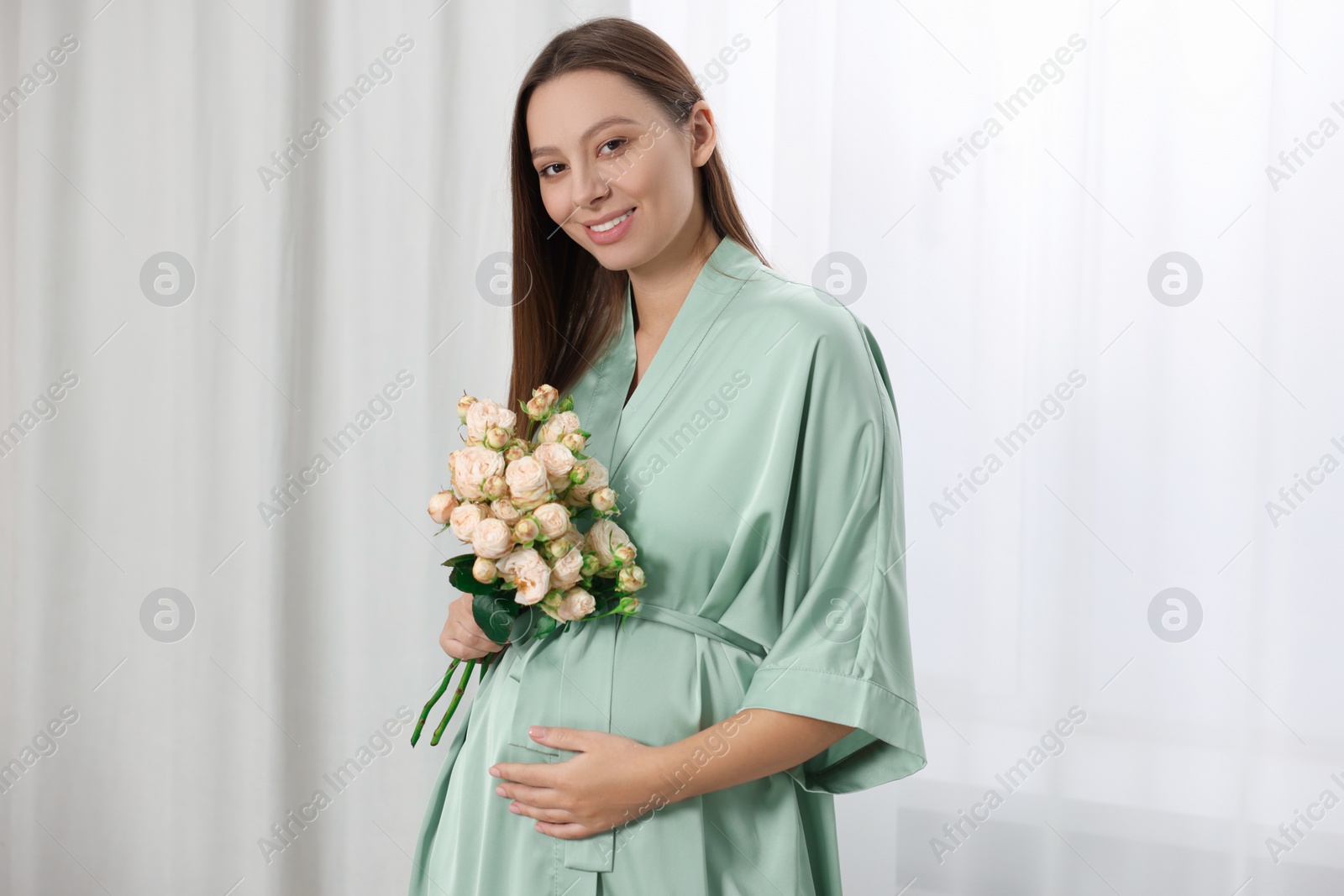 Photo of Beautiful pregnant woman with bouquet of roses near window indoors