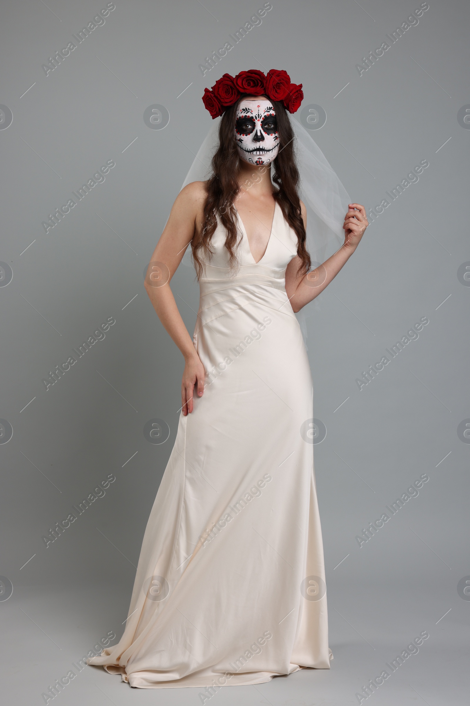 Photo of Young woman in scary bride costume with sugar skull makeup and flower crown on light grey background. Halloween celebration