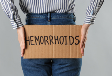 Photo of Woman holding carton sign with word HEMORRHOIDS on light grey background, closeup