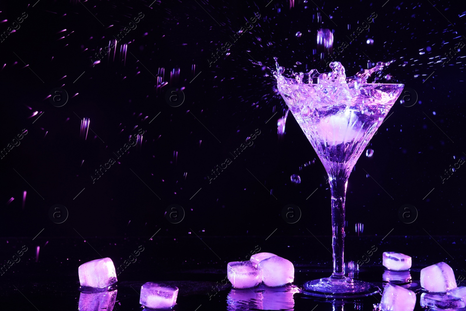 Photo of Glass of martini with ice and splashes in neon lights on dark background. Space for text