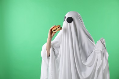 Photo of Creepy ghost. Person in white sheet with tasty hamburger on green background, space for text
