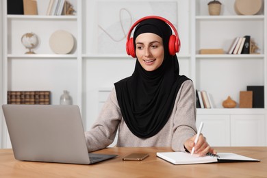 Photo of Muslim woman in headphones writing notes near laptop at wooden table in room