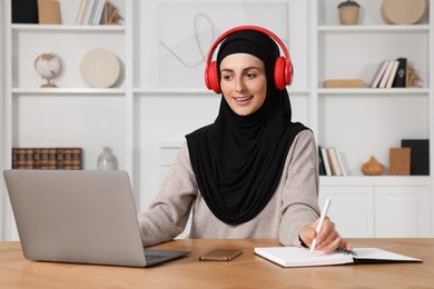 Muslim woman in headphones writing notes near laptop at wooden table in room