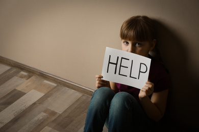 Sad little girl with sign HELP sitting on floor indoors, space for text. Child in danger