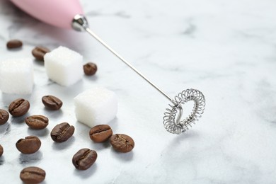 Pink milk frother wand, sugar cubes and coffee beans on white marble table, closeup