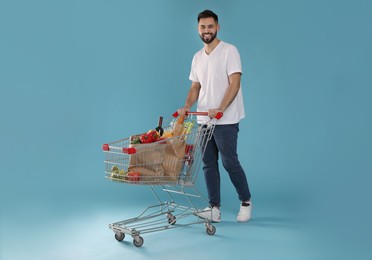 Photo of Happy man with shopping cart full of groceries on light blue background