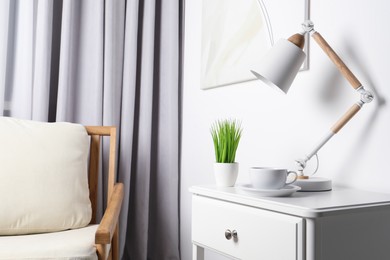 Stylish lamp, houseplant and cup of drink on white side table in light room. Interior design
