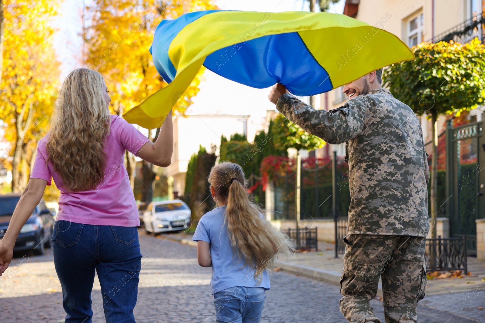 Photo of Soldier in military uniform with his family running and holding Ukrainian flag on city street, back view