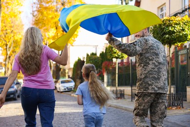 Photo of Soldier in military uniform with his family running and holding Ukrainian flag on city street, back view