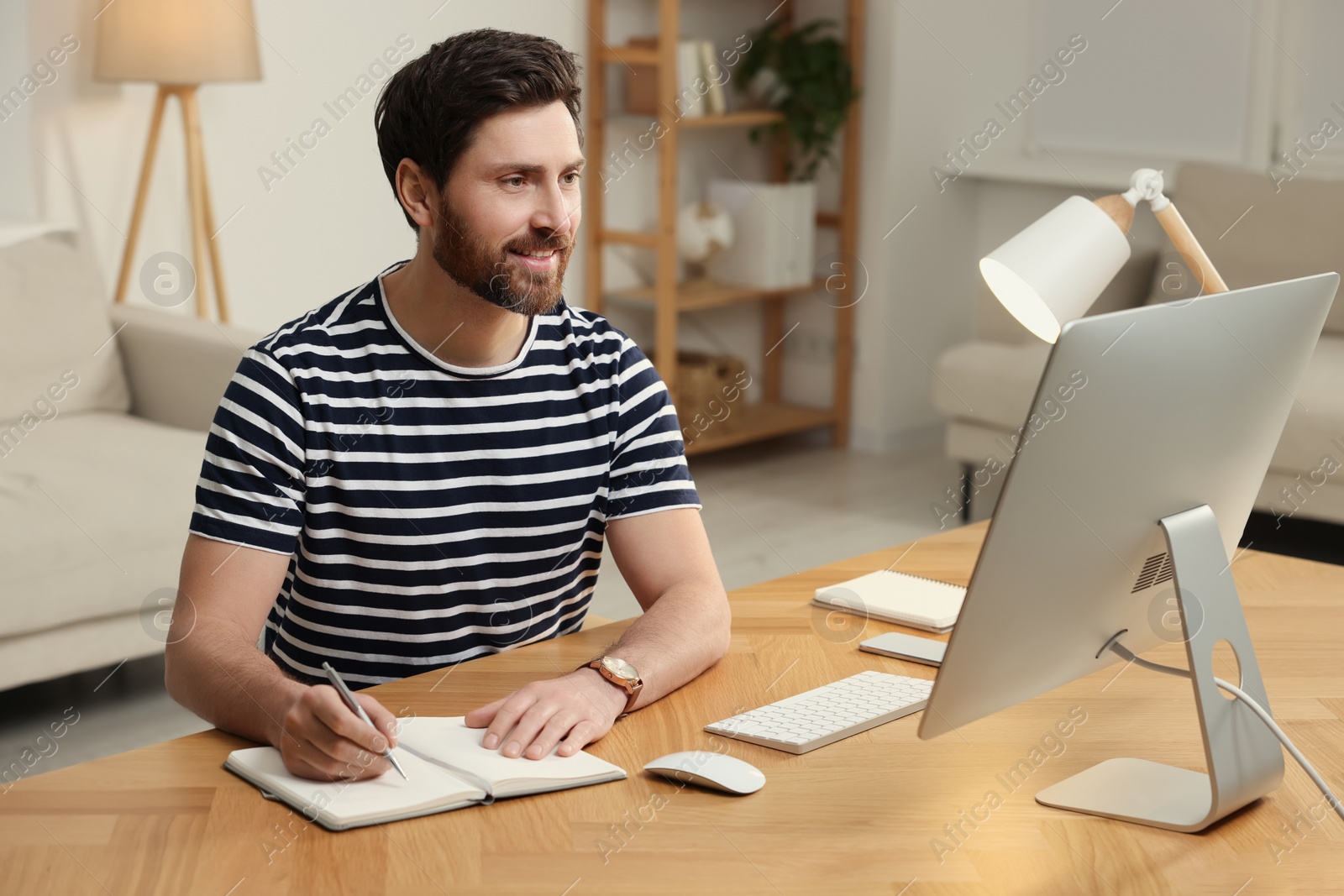 Photo of Home workplace. Happy man taking notes while working with computer at wooden desk in room