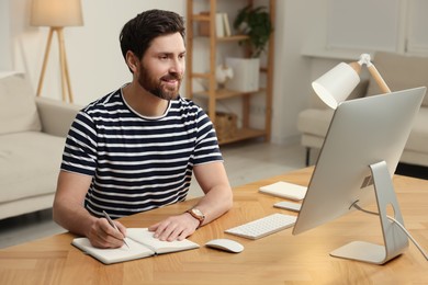 Home workplace. Happy man taking notes while working with computer at wooden desk in room