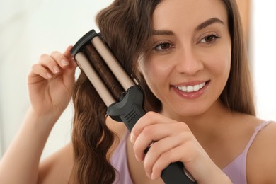 Young woman using modern curling iron indoors
