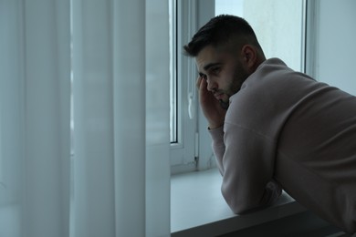 Photo of Sad man near window at home. Space for text