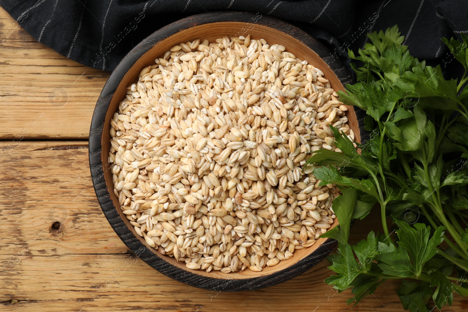 Photo of Dry pearl barley in bowl and parsley on wooden table, top view