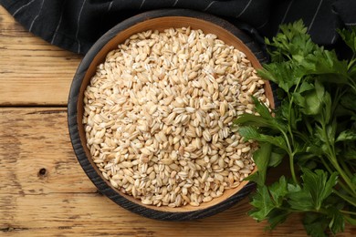 Dry pearl barley in bowl and parsley on wooden table, top view