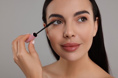 Beautiful young woman applying mascara on grey background, closeup