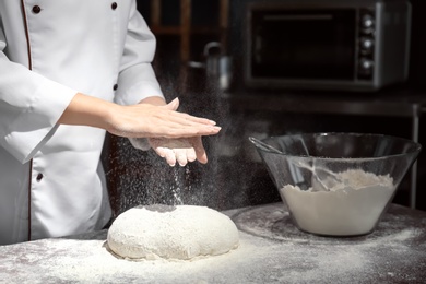 Photo of Female chef sprinkling flour over dough on table in kitchen