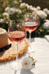 Photo of Glasses of rose wine, straw hat and beautiful flower on white wooden table outdoors