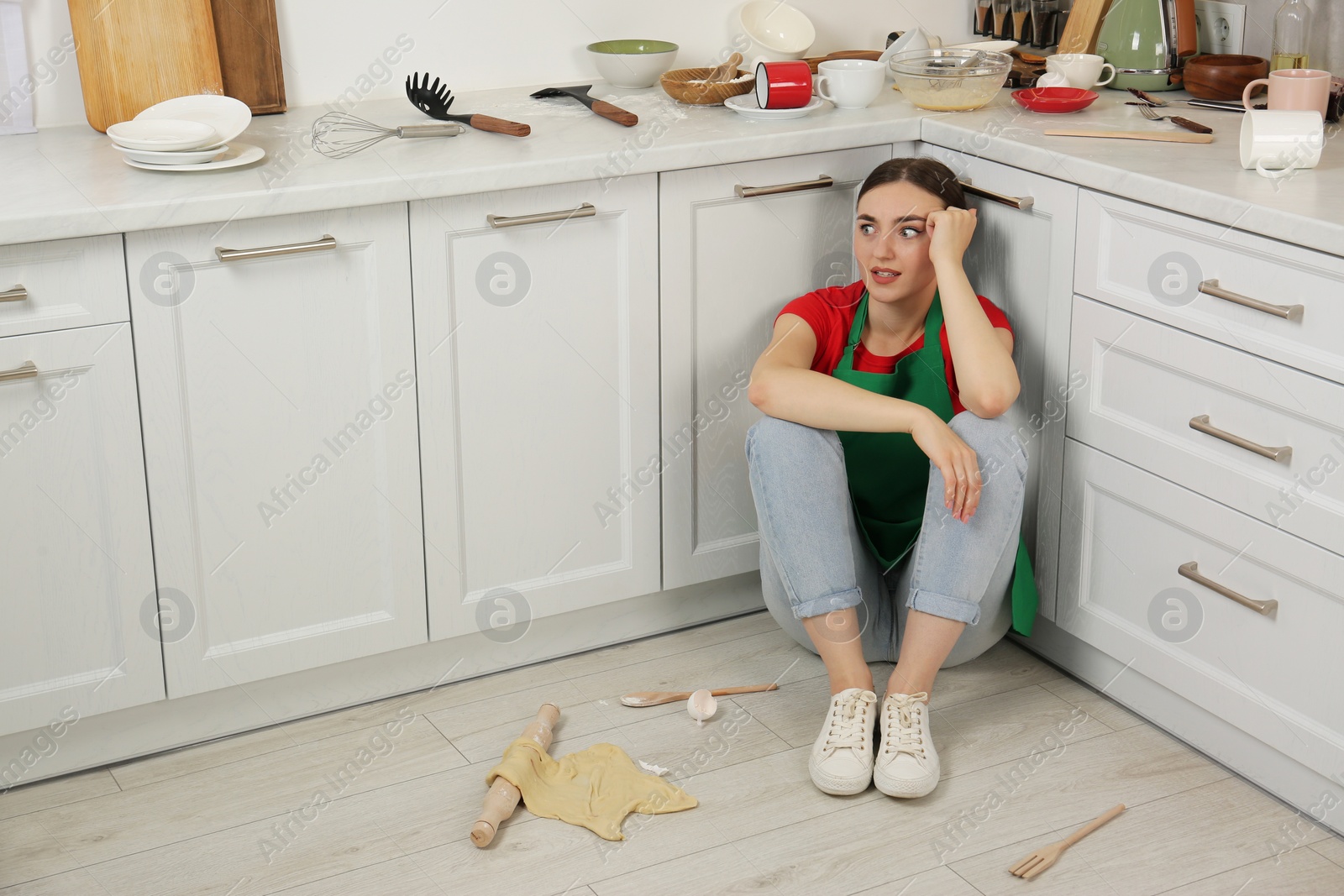 Photo of Tired woman sitting on dirty floor with utensils and food leftovers in messy kitchen