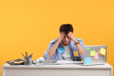 Tired young man working at white table on orange background. Deadline concept