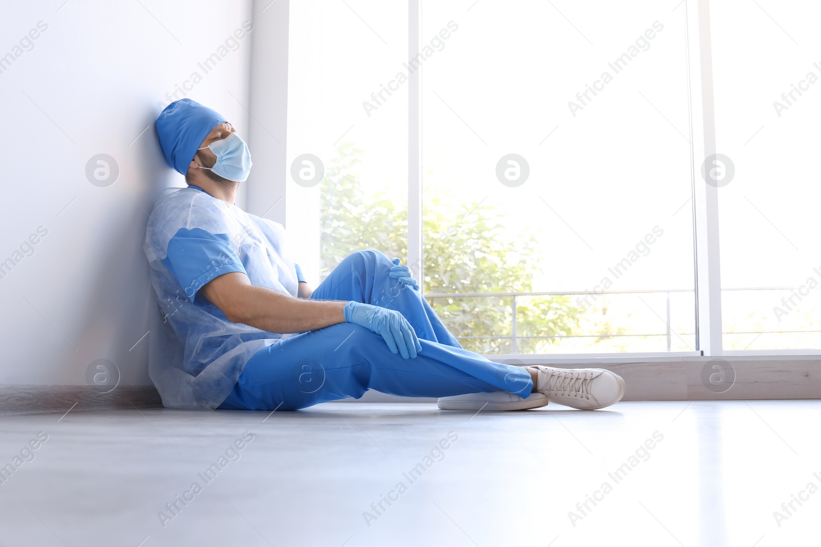 Photo of Exhausted doctor sitting on floor indoors. Stress of health care workers during COVID-19 pandemic