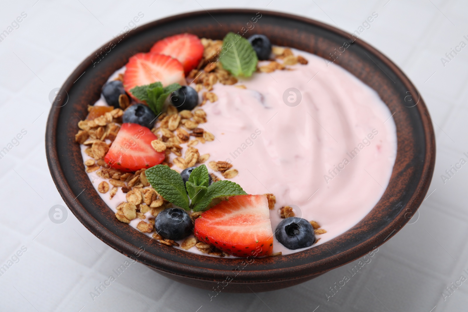Photo of Bowl with yogurt, berries and granola on white tiled table, closeup