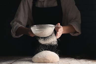 Photo of Woman sprinkling flour over dough at wooden table on dark background, closeup