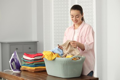 Photo of Young woman with basket full of clean laundry at wooden table indoors
