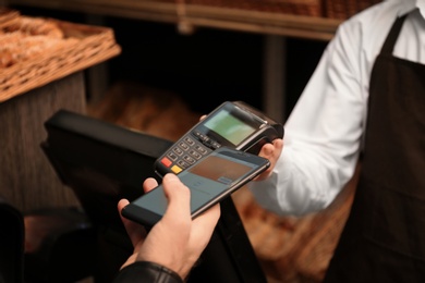 Photo of Man with smartphone using payment terminal at shop, closeup