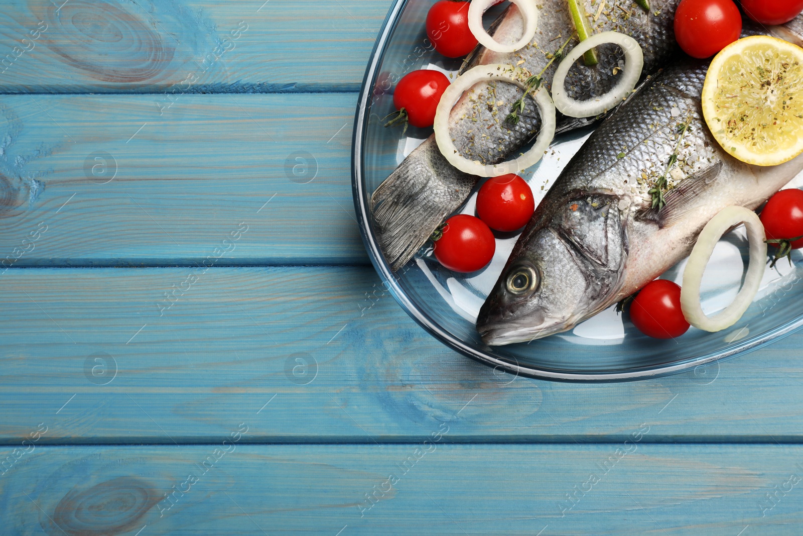 Photo of Glass baking tray with sea bass fish and ingredients on light blue wooden table, top view. Space for text