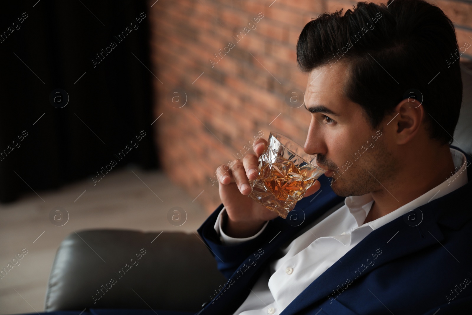 Photo of Young man with glass of whiskey near brick wall indoors. Space for text