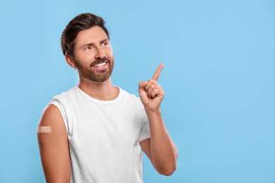 Photo of Man with sticking plaster on arm after vaccination pointing at something against light blue background, space for text