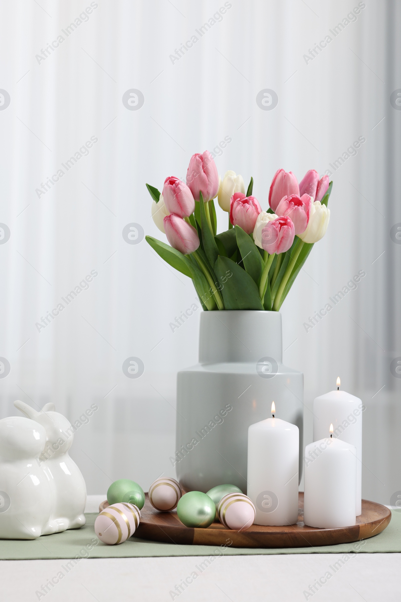 Photo of Easter decorations. Bouquet of tulips, painted eggs, burning candles and bunny figures on white table indoors