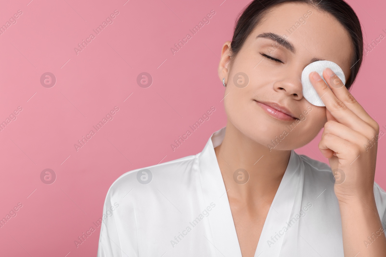 Photo of Young woman cleaning her face with cotton pad on pink background. Space for text