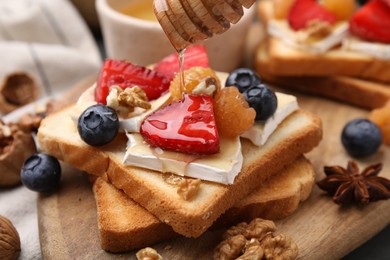 Pouring honey onto tasty sandwiches with brie cheese, berries and walnuts on table, closeup