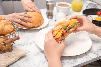 Photo of Woman holding tasty croissant sandwich over plate at table