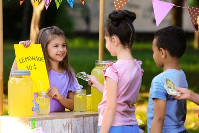 Photo of Little girl selling natural lemonade to kids in park. Summer refreshing drink