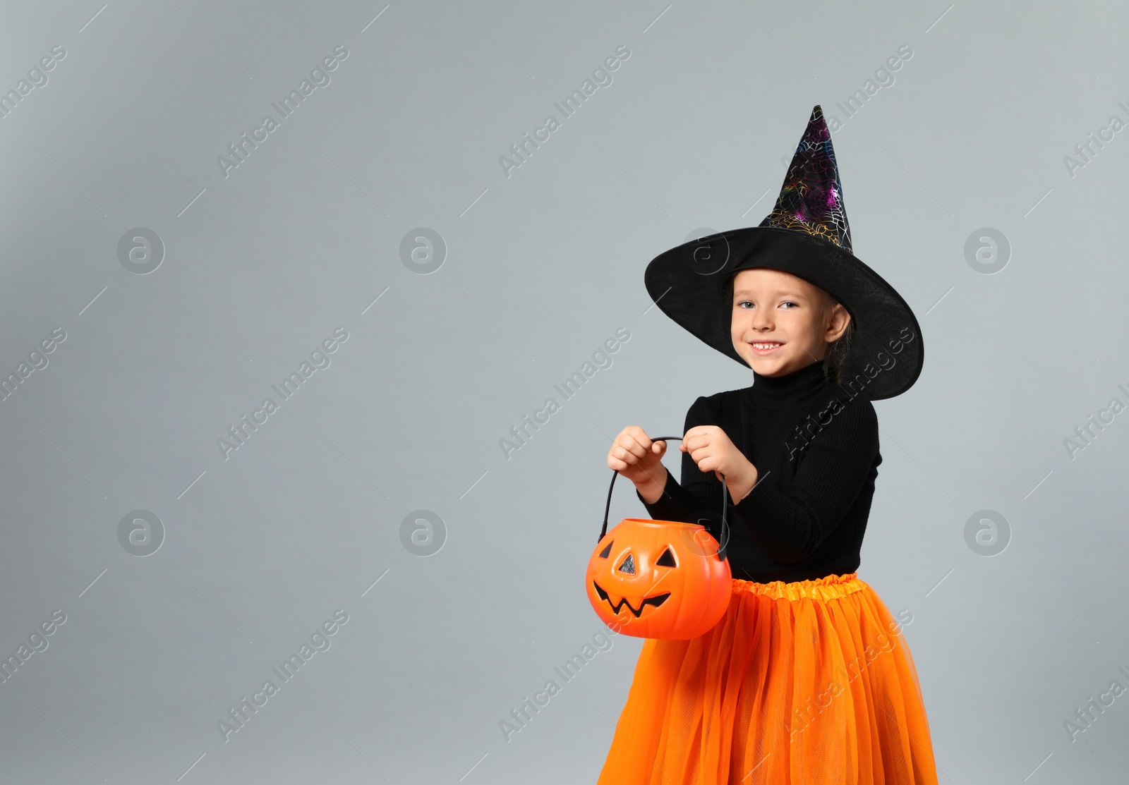 Photo of Cute little girl with pumpkin candy bucket wearing Halloween costume on grey background, space for text