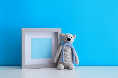 Photo of Empty photo frame and toy bear on white table near light blue wall. Space for design