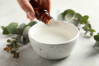 Woman dripping eucalyptus essential oil from bottle into bowl at white table, closeup