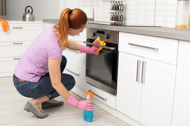 Woman cleaning modern oven with rag and detergent in kitchen