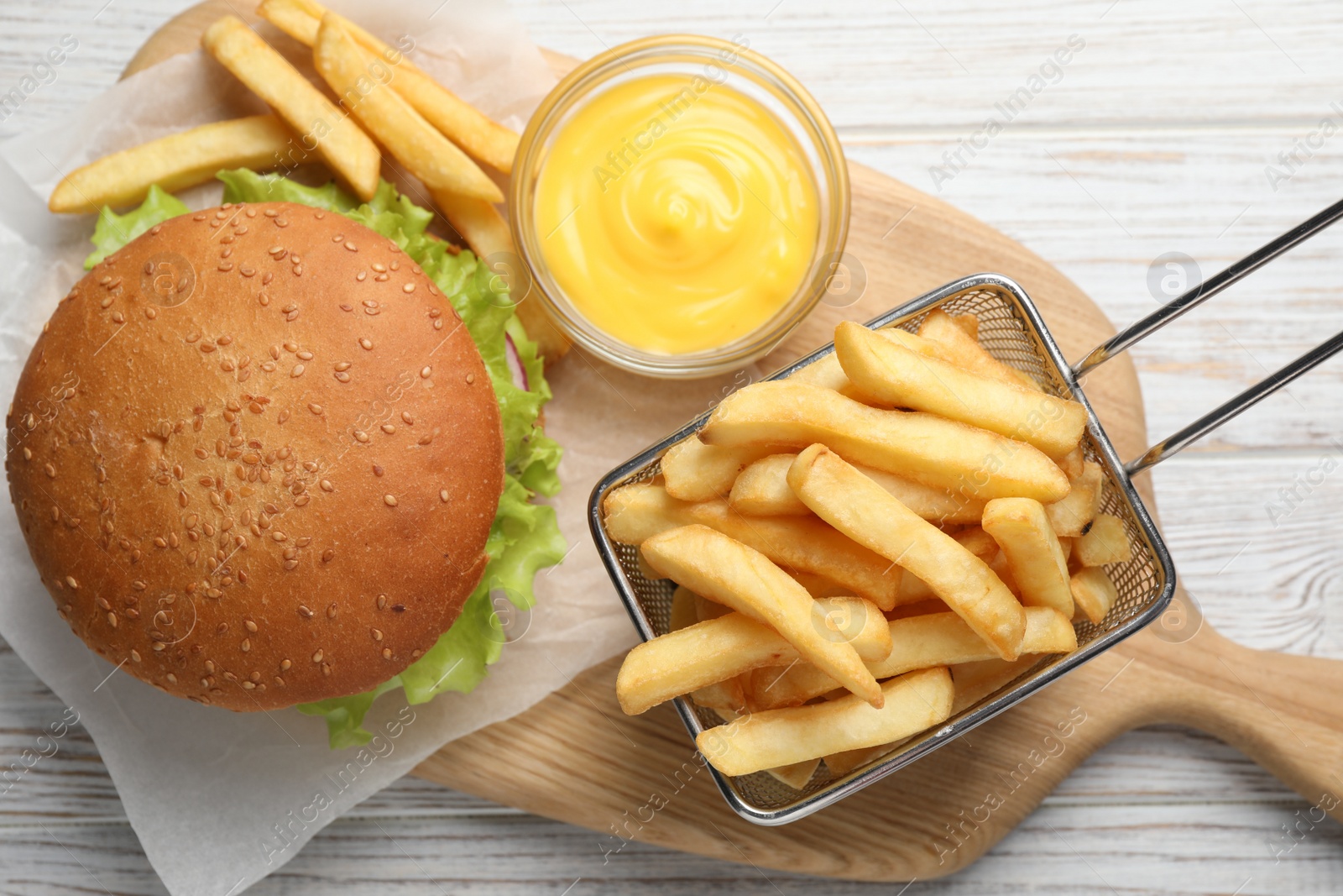 Photo of French fries in frying basket, tasty burger and sauce on white wooden table, flat lay