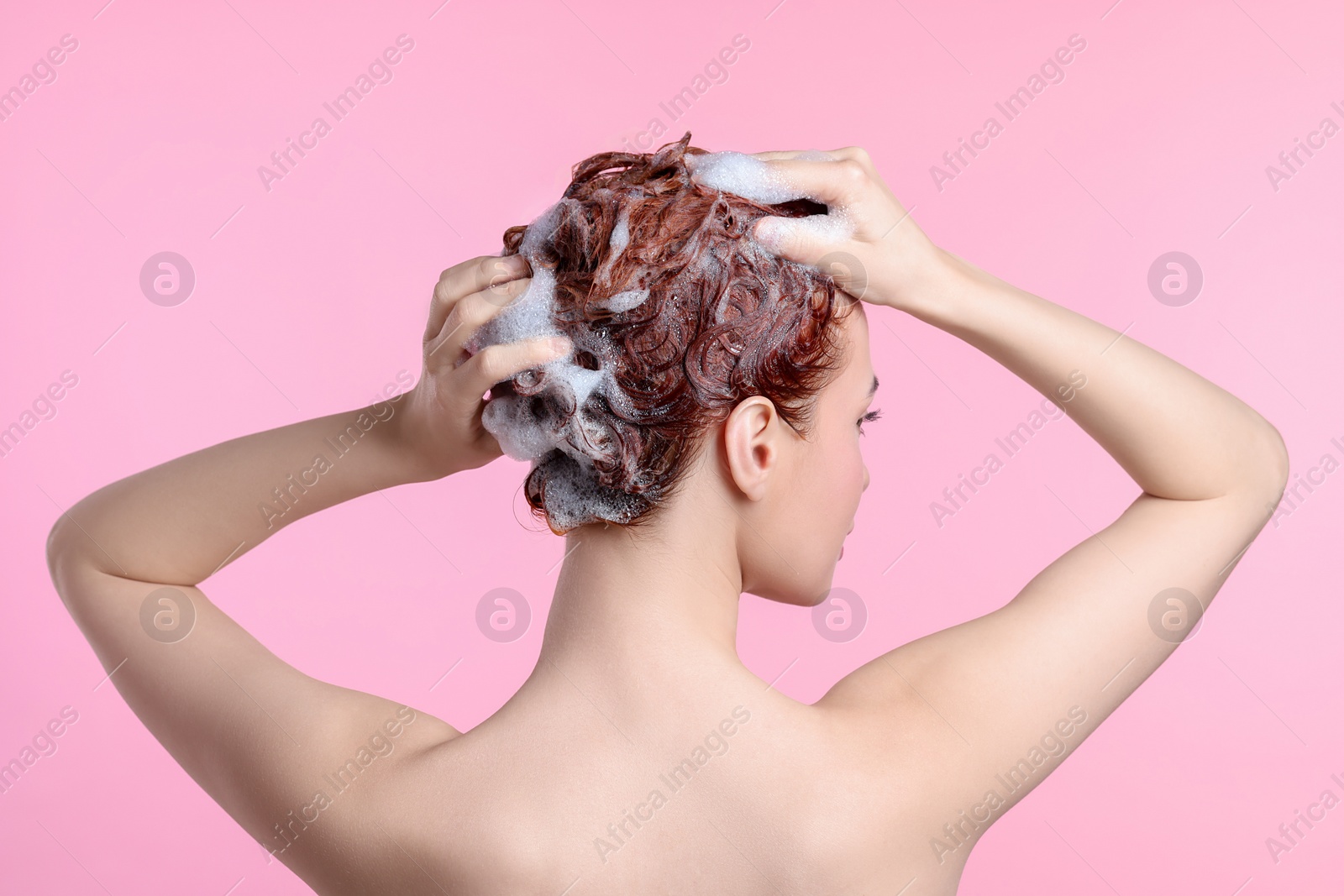Photo of Young woman washing her hair with shampoo on pink background, back view