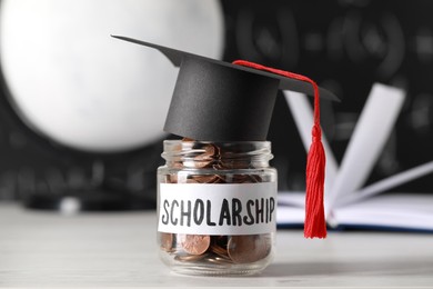 Photo of Scholarship concept. Glass jar with coins and graduation cap on white wooden table, closeup