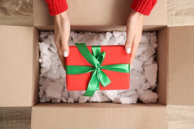 Woman putting Christmas gift box into parcel at wooden table, top view. Sending present by mail